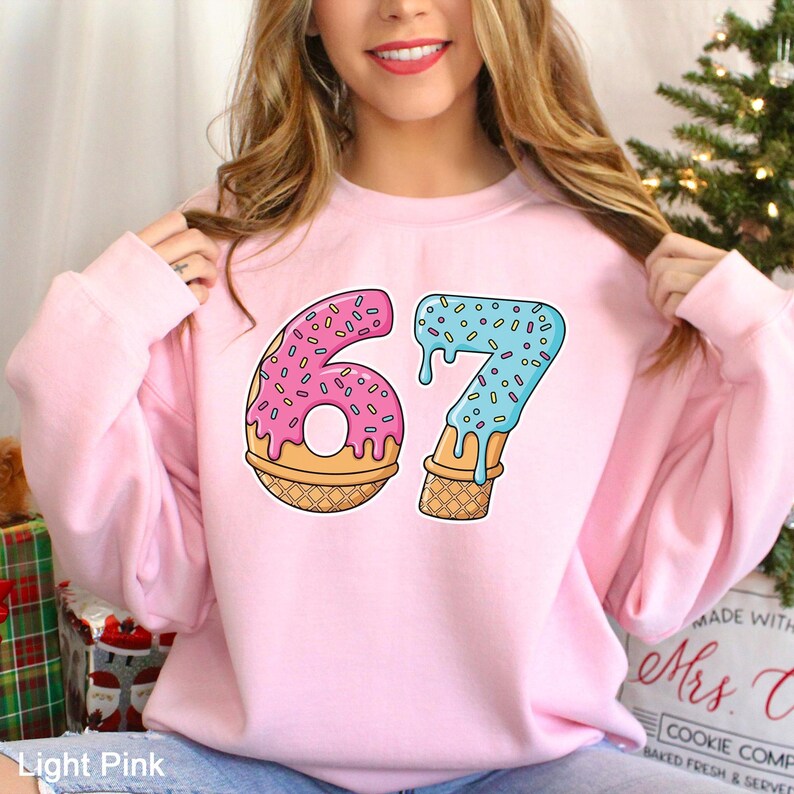 May include: Light pink sweatshirt featuring the number 67 designed as a donut and ice cream cone. The number 6 is a pink donut with sprinkles, and the 7 is a blue ice cream cone with sprinkles. The sweatshirt is a light pink color.