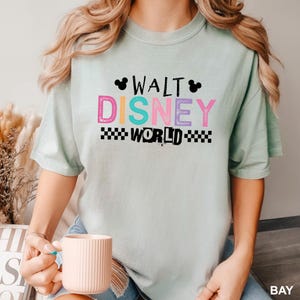 May include: A light green short-sleeve t-shirt featuring the text WALT DISNEY WORLD in colorful block letters. The design includes two small black Mickey Mouse head silhouettes and a black and white checkered pattern below the word WORLD.