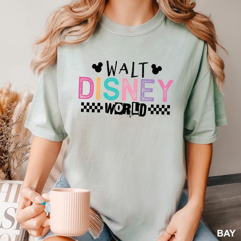 May include: A light green short-sleeve t-shirt featuring the text WALT DISNEY WORLD in colorful block letters. The design includes two small black Mickey Mouse head silhouettes and a black and white checkered pattern below the word WORLD.