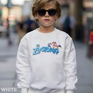 Puede incluir: Sudadera blanca con la palabra "Zootopia" en azul y personajes de dibujos animados. La sudadera es de cuello redondo. La palabra "WHITE" est&aacute; en la parte inferior de la imagen.