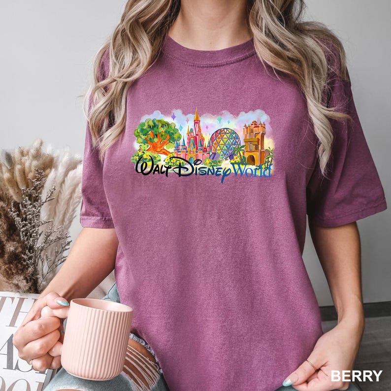 K&ouml;nnte beinhalten: Ein beerenfarbenes Kurzarm-T-Shirt mit einem farbenfrohen Grafikdruck der Wahrzeichen des Walt Disney World Parks, darunter ein Schloss, eine geod&auml;tische Kuppel und ein gro&szlig;er Baum, mit dem in Schwarz gedruckten Text Walt Disney World.