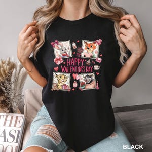 May include: Black t-shirt featuring four framed cartoon characters with Valentine's Day themed elements. The words "HAPPY VALENTINE'S DAY" are in pink. The shirt is made of a soft material.