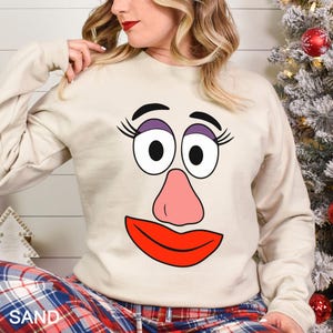 May include: Sand-colored sweatshirt featuring a cartoon face with large eyes, a pink nose, and a red mouth. The sweatshirt has long sleeves and is paired with plaid pajama pants. The word "SAND" is printed at the bottom.