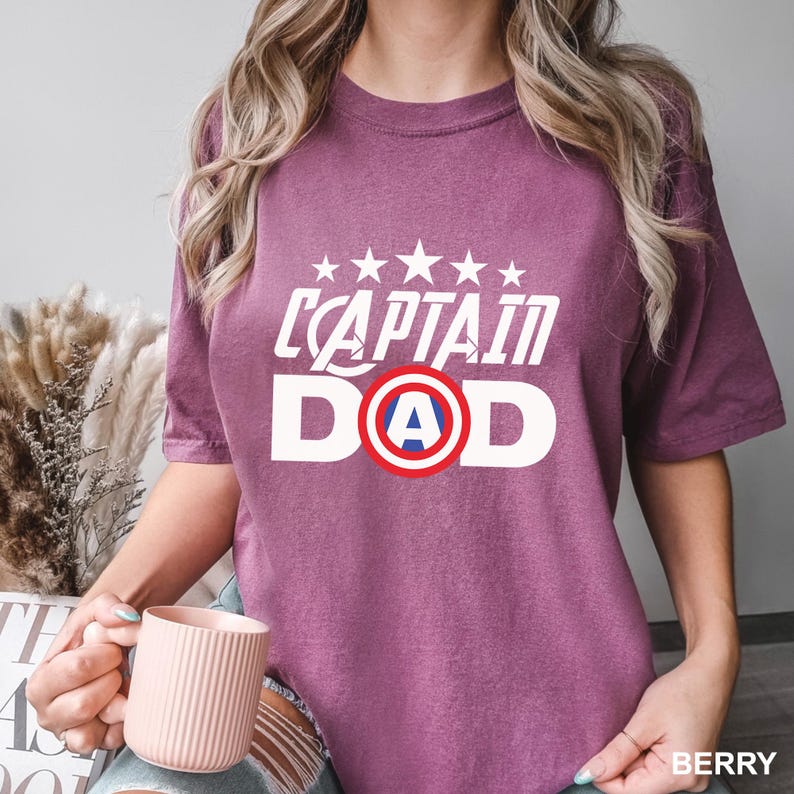 Puede incluir: Camiseta color baya con el texto blanco "CAPTAIN DAD", con un dise&ntilde;o de escudo con la letra "A" en el centro. Encima del texto hay cinco estrellas blancas. La camiseta es de cuello redondo y manga corta.