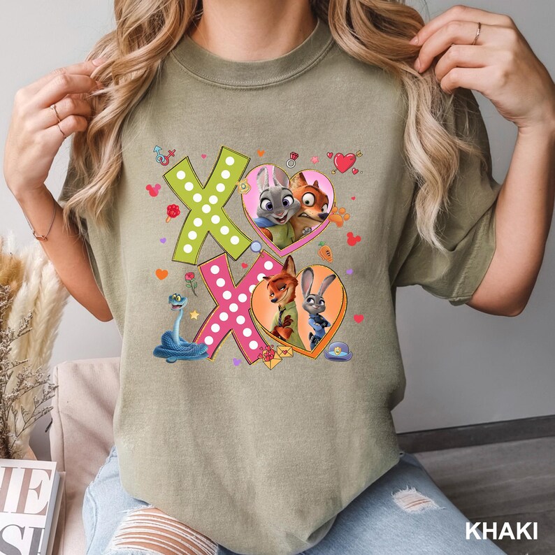 May include: Khaki-colored t-shirt featuring the characters from Zootopia. The design includes the letters "XO" with heart-shaped frames containing images of the characters. Additional graphics include hearts, flowers, and a blue snake.