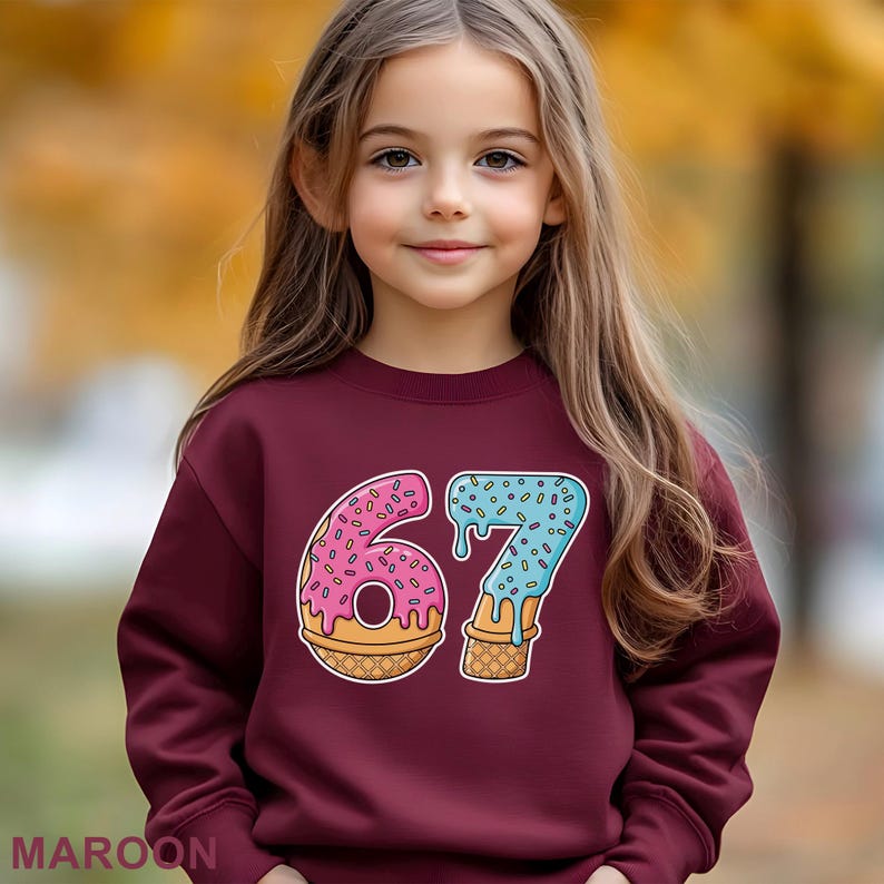 May include: A maroon sweatshirt with a colorful graphic of the number "67" designed as a donut and ice cream cone. The donut is pink with sprinkles, and the ice cream cone is blue with sprinkles. The word "MAROON" is printed at the bottom.