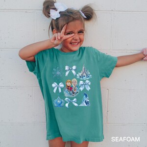 May include: A teal-colored t-shirt featuring a graphic of characters and elements from a popular animated film. The design includes images of a castle, snowflakes, bows, and characters. The text "SEAFOAM" is printed at the bottom of the shirt.
