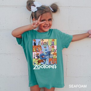May include: A teal-colored t-shirt featuring a colorful graphic from the animated film "Zootopia." The graphic includes various animal characters in a grid pattern, with the film's title in white block letters. The shirt is labeled "SEAFOAM" at the bottom.