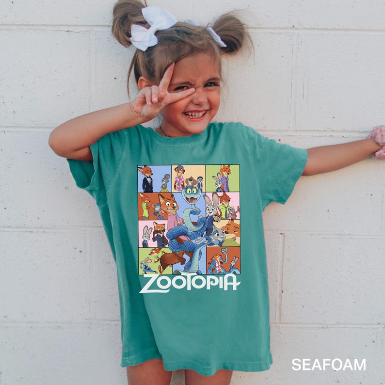 May include: A teal-colored t-shirt featuring a colorful graphic from the animated film "Zootopia." The graphic includes various animal characters in a grid pattern, with the film's title in white block letters. The shirt is labeled "SEAFOAM" at the bottom.