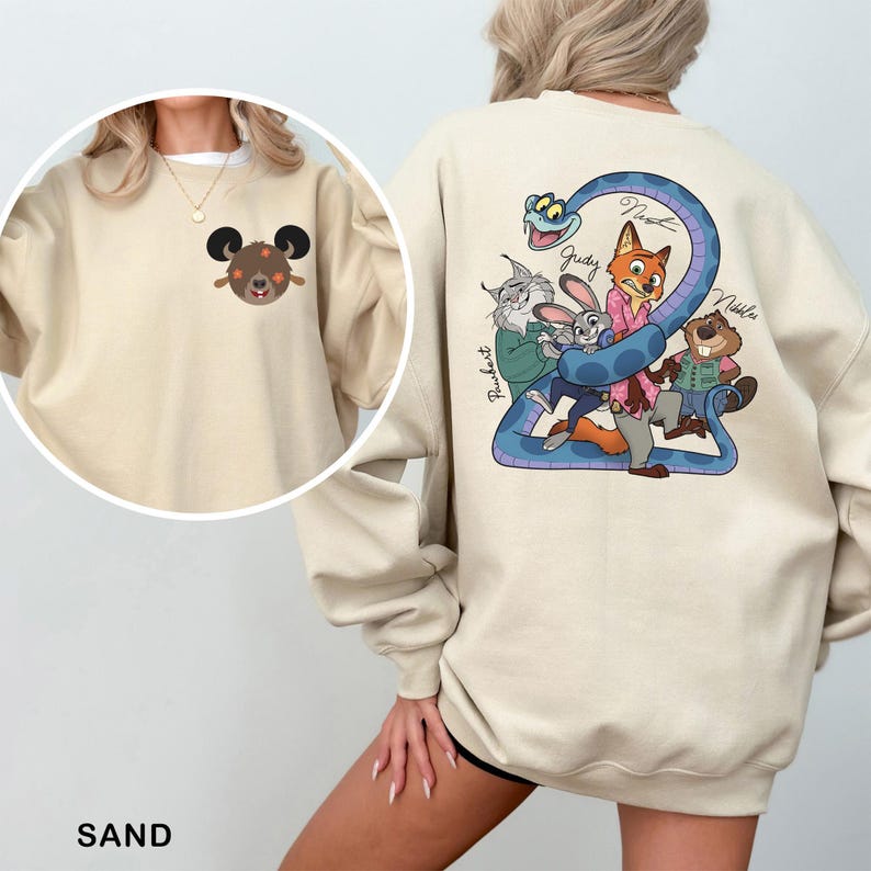 Op de afbeelding: Zandkleurige sweatshirt met een cartoonafbeelding op de rug met een blauwe slang en verschillende dierenfiguren. De voorkant van de sweatshirt heeft een kleine cartoon berenafbeelding. Het woord "SAND" staat onderaan gedrukt.