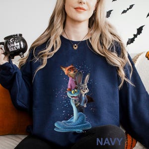 K&ouml;nnte beinhalten: Marineblaues Sweatshirt mit einer farbenfrohen Grafik von Zeichentrickfiguren. Das Design zeigt einen Fuchs in einem rosa Hemd, ein Kaninchen in einem blauen Anzug und eine blaue Schlange. Das Wort "NAVY" ist unten aufgedruckt.