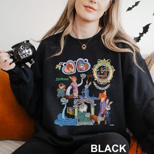 May include: Black sweatshirt with a colourful graphic of characters from the animated film "Zootopia." The design includes images of Nick Wilde, Judy Hopps, and other characters in a framed design. The word "Zootopia" is printed in green. The word "BLACK" is printed at the bottom.