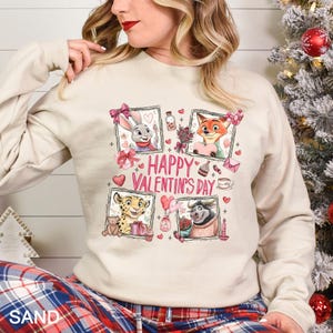 May include: Sand-colored sweatshirt featuring a Valentine's Day design. The graphic includes cartoon characters in framed images, hearts, and the text "Happy Valentine's Day." The sweatshirt has long sleeves and a crew neck.