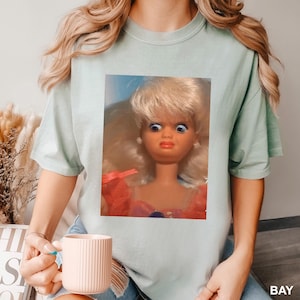May include: A light green t-shirt featuring a close-up image of a vintage doll with blonde hair, blue eyes, and a surprised expression. The doll is wearing a pink and red dress. The word "BAY" is visible in the bottom right corner.