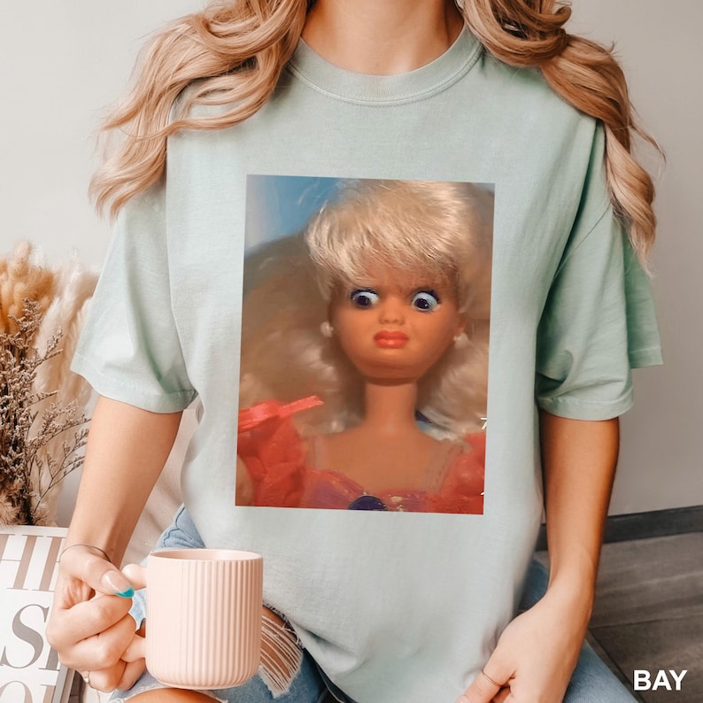 May include: A light green t-shirt featuring a close-up image of a vintage doll with blonde hair, blue eyes, and a surprised expression. The doll is wearing a pink and red dress. The word "BAY" is visible in the bottom right corner.