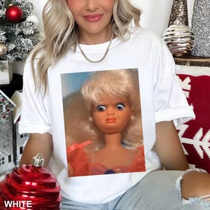 May include: White t-shirt featuring a close-up of a vintage doll with a surprised expression. The doll has blonde hair, blue eyes, and is wearing a pink dress. The word "WHITE" is printed at the bottom of the image.