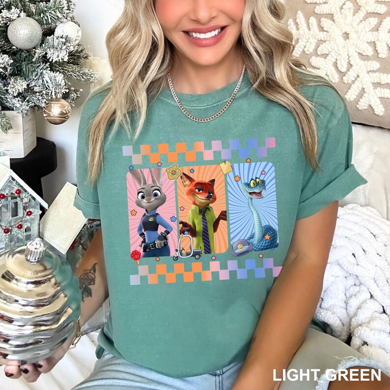 May include: A light green short-sleeve t-shirt featuring a graphic print of three animated characters: a rabbit in a police uniform, a fox in a green shirt and striped tie, and a blue snake. The design includes a checkered border and stars.