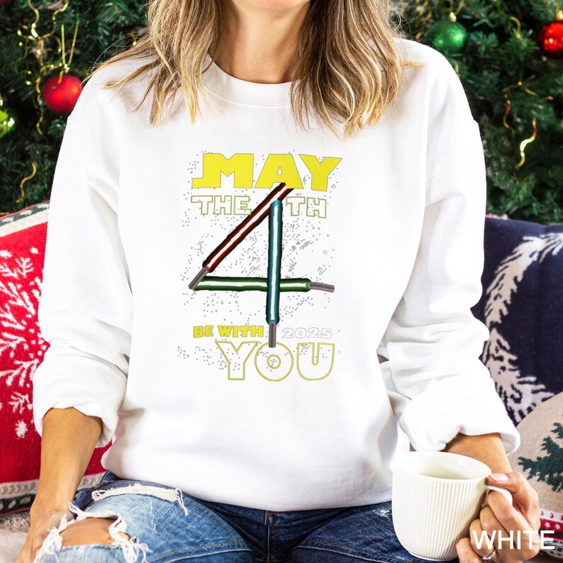 May include: White sweatshirt featuring the text "MAY THE 4TH BE WITH YOU 2025" in yellow and green. The design includes a stylized number 4 with lightsaber-like lines. The person is holding a white mug.