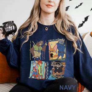 May include: Navy blue sweatshirt featuring a colorful graphic of characters from the animated movie Zootopia. The design includes a sloth, fox, buffalo, rabbit, and raccoon. The word "NAVY" is printed at the bottom of the sweatshirt. A black mug with the words "Witches Brew" is held in the hand.
