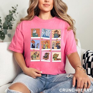 May include: A pink t-shirt featuring a grid of cartoon character images. The images are in a polaroid style with a star at the bottom. The text "CRUNCHBERRY" is at the bottom right of the shirt.