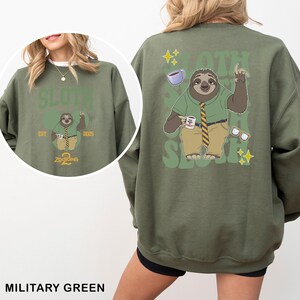 K&ouml;nnte beinhalten: Milit&auml;rgr&uuml;nes Sweatshirt mit einem Cartoon-Faultier-Charakter aus Zootopia. Auf der Vorderseite befindet sich ein kreisf&ouml;rmiges Design mit dem Text "SLOTH 2 Zootopia 2025". Auf der R&uuml;ckseite ist eine gr&ouml;&szlig;ere Faultier-Illustration mit Kaffee, Brille und dem Text "SLOTH" zu sehen.