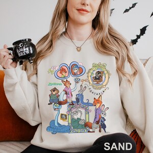 May include: A sand-coloured sweatshirt with a colourful graphic of characters from the animated film "Zootopia." The design includes various animal characters in detailed illustrations, with the word "Zootopia" displayed. The word "SAND" is printed at the bottom.