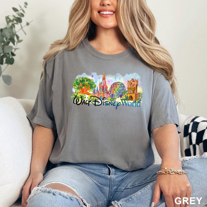 K&ouml;nnte beinhalten: Ein graues Kurzarm-T-Shirt mit einem farbenfrohen Grafikdruck der Wahrzeichen des Walt Disney World Parks, darunter ein Baum, ein Schloss, eine geod&auml;tische Kuppel und ein Turm, mit dem in blauer Schreibschrift gedruckten Text Walt Disney World.