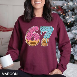 May include: Maroon sweatshirt featuring a playful graphic of the number 67 designed as a donut and ice cream cone. The number 6 is a pink donut with sprinkles, and the 7 is a blue ice cream cone with sprinkles. The word "MAROON" is at the bottom.