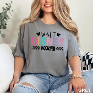 May include: A grey short-sleeve t-shirt featuring the text WALT DISNEY WORLD in a multi-colored graphic design. The word WALT is in black, DISNEY is in pink, blue, and purple, and WORLD is in black with a checkered pattern on both sides.