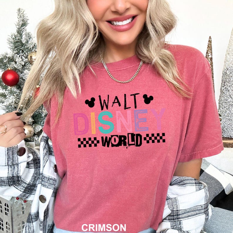 May include: A crimson short-sleeved t-shirt featuring the text WALT DISNEY WORLD in black, pink, yellow, teal, and purple lettering. The design includes two black Mickey Mouse ear silhouettes and black checkered patterns on the sides.