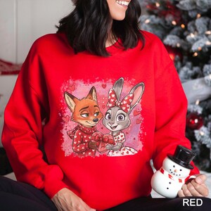 K&ouml;nnte beinhalten: Ein rotes Sweatshirt mit einem Cartoon-Fuchs und -Hasen, die ein herzf&ouml;rmiges Geschenk halten. Der Fuchs tr&auml;gt einen Schal mit Herzmustern, und der Hase hat eine Schleife mit Herzen. Das Wort "RED" ist unten aufgedruckt.