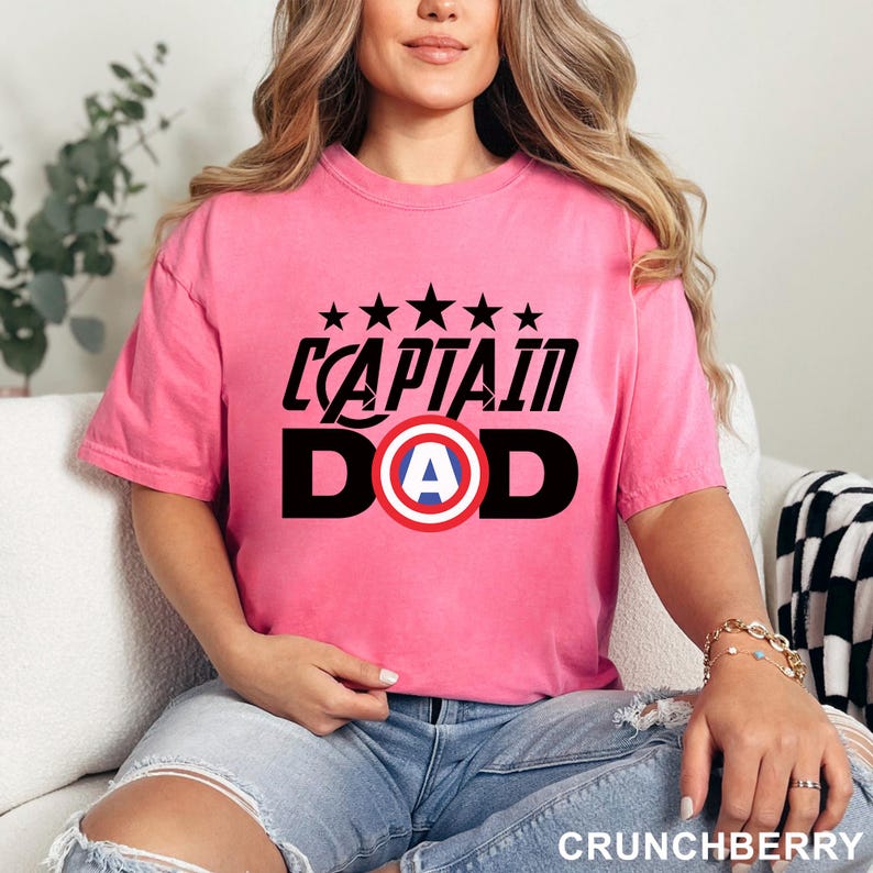 Puede incluir: Una camiseta rosa con el texto "CAPTAIN DAD" en negro, con un dise&ntilde;o de escudo con la letra "A" en el centro. Encima del texto hay cinco estrellas negras. La camiseta la lleva una persona con vaqueros azules.