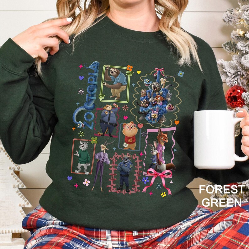 Puede incluir: Sudadera verde bosque con un dise&ntilde;o gr&aacute;fico colorido de personajes de la pel&iacute;cula animada Zootopia. El dise&ntilde;o incluye ilustraciones enmarcadas de personajes de animales, con la palabra "Zootopia". Una taza blanca se sostiene en la mano.