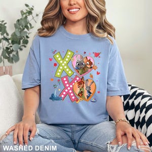 May include: A light blue t-shirt with a graphic featuring the characters from Zootopia. The design includes the letters X and O, with images of Judy Hopps, Nick Wilde, and other characters inside hearts. The words "WASHED DENIM" are printed at the bottom.