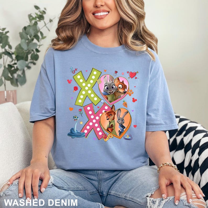 May include: A light blue t-shirt with a graphic featuring the characters from Zootopia. The design includes the letters X and O, with images of Judy Hopps, Nick Wilde, and other characters inside hearts. The words "WASHED DENIM" are printed at the bottom.