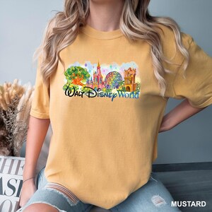 K&ouml;nnte beinhalten: Ein senfgelbes Kurzarm-T-Shirt mit einem farbenfrohen Grafikdruck der Wahrzeichen des Walt Disney World Freizeitparks, einschlie&szlig;lich Schloss und geod&auml;tischer Kuppel, sowie dem Schriftzug Walt Disney World in blauer Schreibschrift.