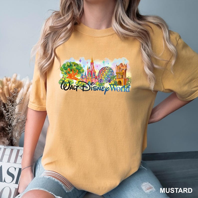 K&ouml;nnte beinhalten: Ein senfgelbes Kurzarm-T-Shirt mit einem farbenfrohen Grafikdruck der Wahrzeichen des Walt Disney World Freizeitparks, einschlie&szlig;lich Schloss und geod&auml;tischer Kuppel, sowie dem Schriftzug Walt Disney World in blauer Schreibschrift.