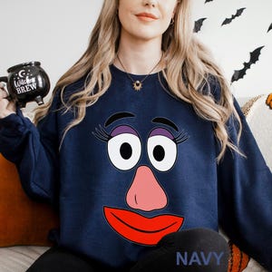 May include: Navy blue sweatshirt featuring a cartoon face with large white eyes, purple eyelashes, a pink nose, and a red mouth. The word "NAVY" is printed at the bottom. The person in the image is holding a black mug that says "Witches Brew".