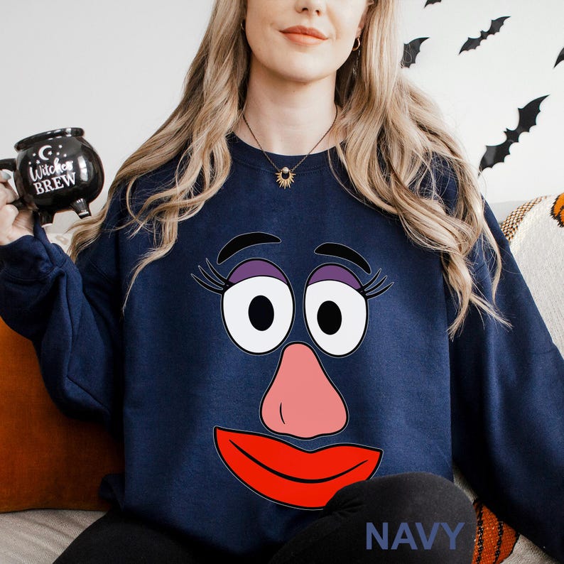 May include: Navy blue sweatshirt featuring a cartoon face with large white eyes, purple eyelashes, a pink nose, and a red mouth. The word "NAVY" is printed at the bottom. The person in the image is holding a black mug that says "Witches Brew".