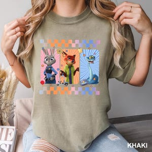 May include: A khaki-colored crew neck t-shirt featuring a graphic print of three animated characters: a rabbit in a police uniform, a fox in a green shirt and tie, and a blue snake. The design includes a checkered border and small star icons.