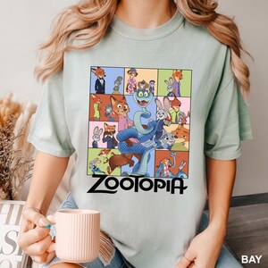 May include: A light green t-shirt featuring a colorful graphic of characters from the animated movie "Zootopia". The design includes a snake, rabbits, foxes, and other animals. The word "Zootopia" is printed in black at the bottom.