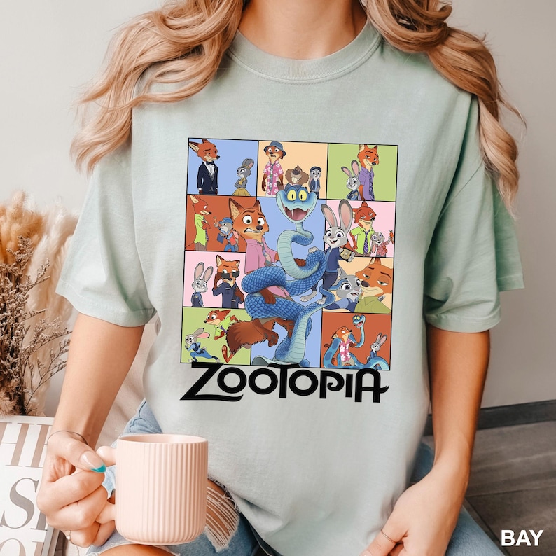 May include: A light green t-shirt featuring a colorful graphic of characters from the animated movie "Zootopia". The design includes a snake, rabbits, foxes, and other animals. The word "Zootopia" is printed in black at the bottom.