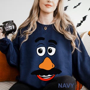 May include: Navy blue sweatshirt featuring a cartoon face design with large white eyes, an orange nose, and a black mustache and eyebrows. The word "NAVY" is printed at the bottom. The person is holding a black mug that says "Witches Brew".