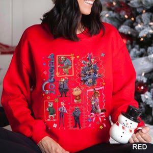 Puede incluir: Una sudadera roja con personajes de la pel&iacute;cula animada "Zootopia". El dise&ntilde;o incluye im&aacute;genes enmarcadas de varios personajes, la palabra "Zootopia" y elementos decorativos. La palabra "RED" est&aacute; impresa en la parte inferior derecha.