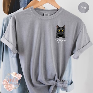 May include: A grey t-shirt with a black cat peeking over the edge of a pocket. The cat has yellow eyes and the text "Name" is printed below the cat.