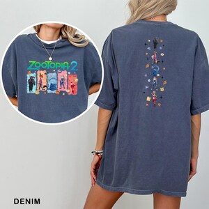 May include: Denim blue t-shirt featuring the text "ZOOTOPIA 2" with colorful character graphics on the front. The back of the shirt has a vertical design with more character illustrations and stars. A casual, oversized fit.