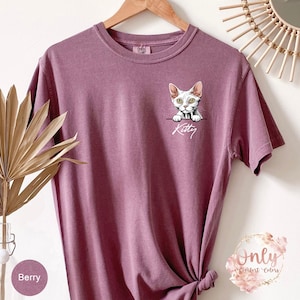May include: A mauve t-shirt with a white cat peeking out of a pocket and the word "Kitty" written below.