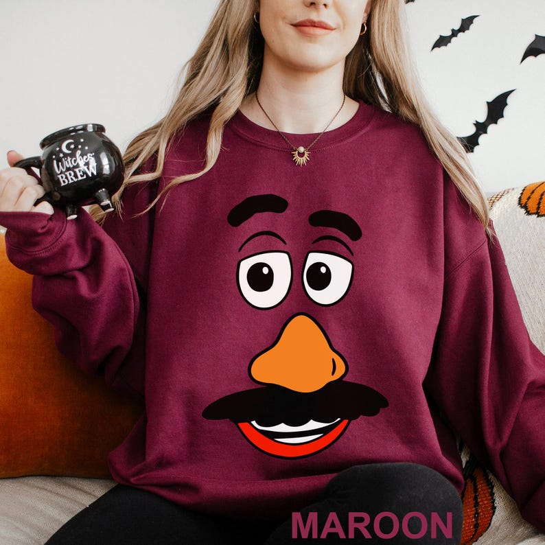 May include: Maroon-colored sweatshirt featuring a cartoon face with large eyes, an orange nose, and a black mustache. The word "MAROON" is printed at the bottom. A black mug with the words "Witches Brew" is held in the left hand.