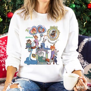 May include: White sweatshirt with a colourful graphic of characters from the animated film "Zootopia." The design includes various animal characters in ornate frames and the word "Zootopia." The sweatshirt is a crew neck style.