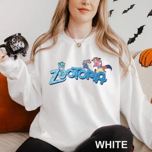 Puede incluir: Sudadera blanca con la palabra "Zootopia" en azul y personajes de dibujos animados. La sudadera tiene mangas largas y cuello redondo. La palabra "WHITE" est&aacute; impresa en la parte inferior de la imagen.